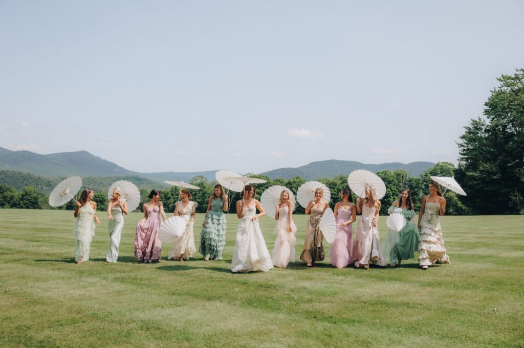 A bride and her bridal party walk barefoot across a grassy field holding white parasols, dressed in flowing pastel gowns with mountains in the background on a sunny day. The vibe is country club meet royalty. Photographed by Lillian Seibert, Vermont Wedding Photographer.  New England Wedding Content Creator.