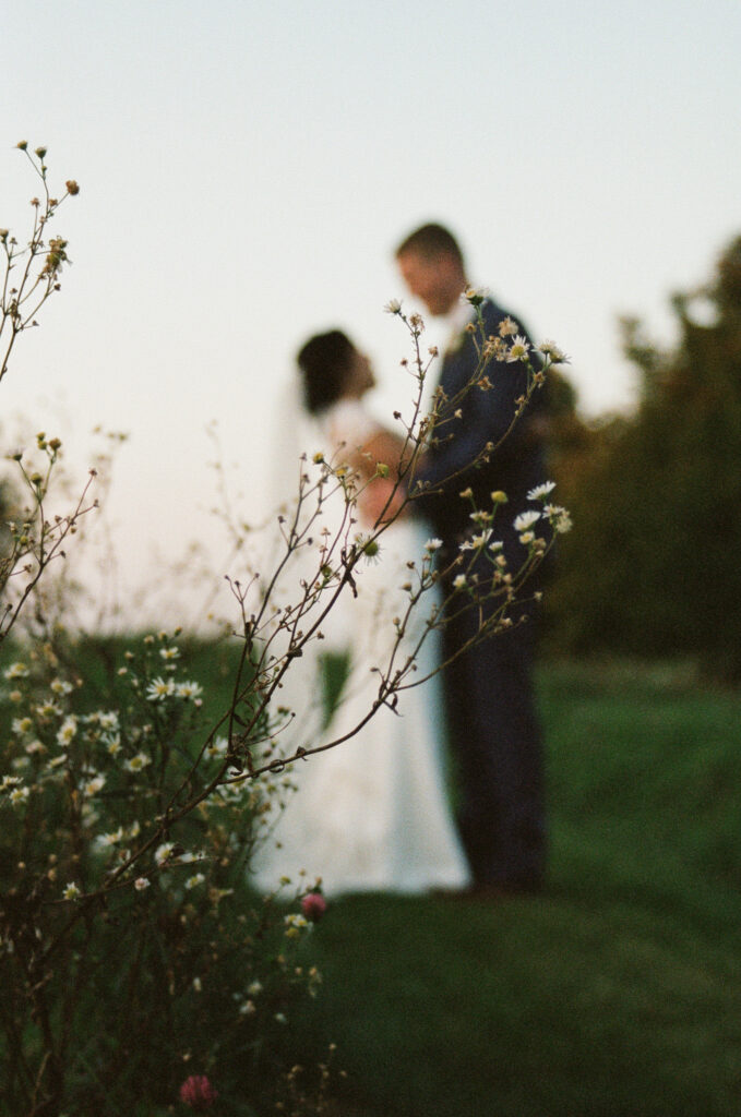 Artistic film wedding photo taken by a Vermont and New England-based wedding photographer, featuring a flower in sharp focus with a softly blurred bride and groom in the background