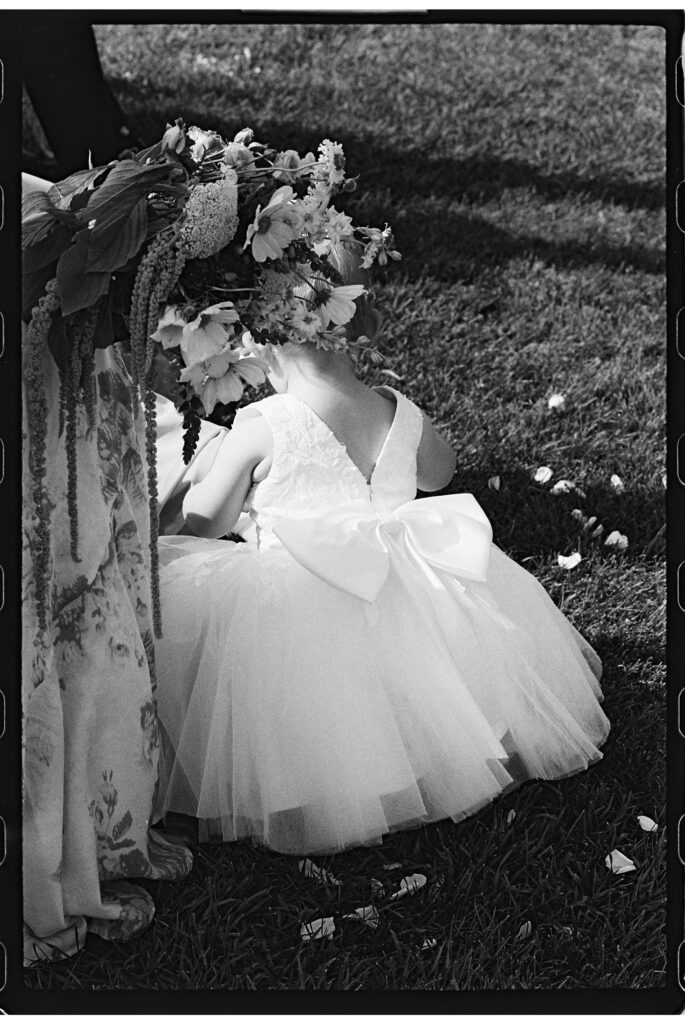 Black and white film photo of a flower girl admiring the bow on the back of her white dress, captured by a Vermont and New England wedding photographer